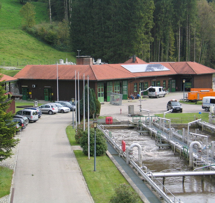 Kläranlage beim AUV-Chiemsee, im Vordergrund ein klärbecken, hinten ein Gebäude mit Solarpanelen, davor stehen Autos und ein Mitarbeiter sitz auf der Bank vor dem Gebäude