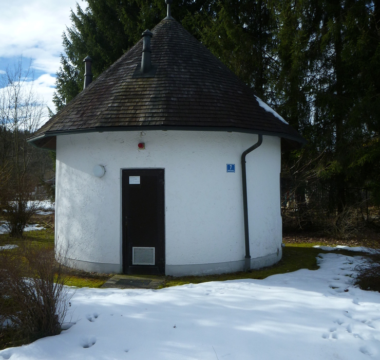 Ortskanal Chieming. Ein weißes kleine Rundes gebäude mit Kegelförmigem Holzdach. Am Gebäude rechts ein blaues Schild mit der Zahl 7. Vor dem Gebäude liegt noch Schnee.