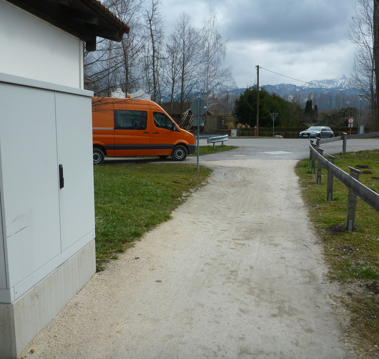 Ortskanal Betreuung Gemeinde Übersee. Ein geschotterer Weg. Rechts durch einen Holzzaun begrenzt, Links ein Stromverteilerkasten. Ebenfalls auf der Linken Seite weiter vorne ein Roter Kastenwagen der Stadt